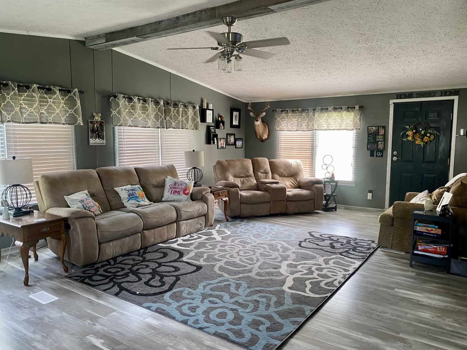 A cozy living room with beige reclining sofas along two walls. There's a grey and white floral patterned rug on the light wooden floor. The walls are adorned with family photos and a mounted deer head. The ceiling has exposed wooden beams and a ceiling fan.