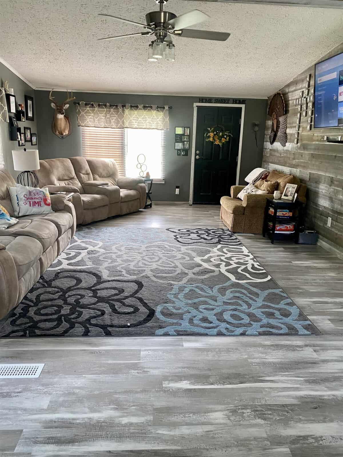 A cozy living room with a large patterned rug and gray-toned laminate flooring. It features plush beige sofas, wall-mounted TV, decorative accents including a deer head, picture frames, and an entry door adorned with a wreath. A ceiling fan hangs from the textured ceiling.