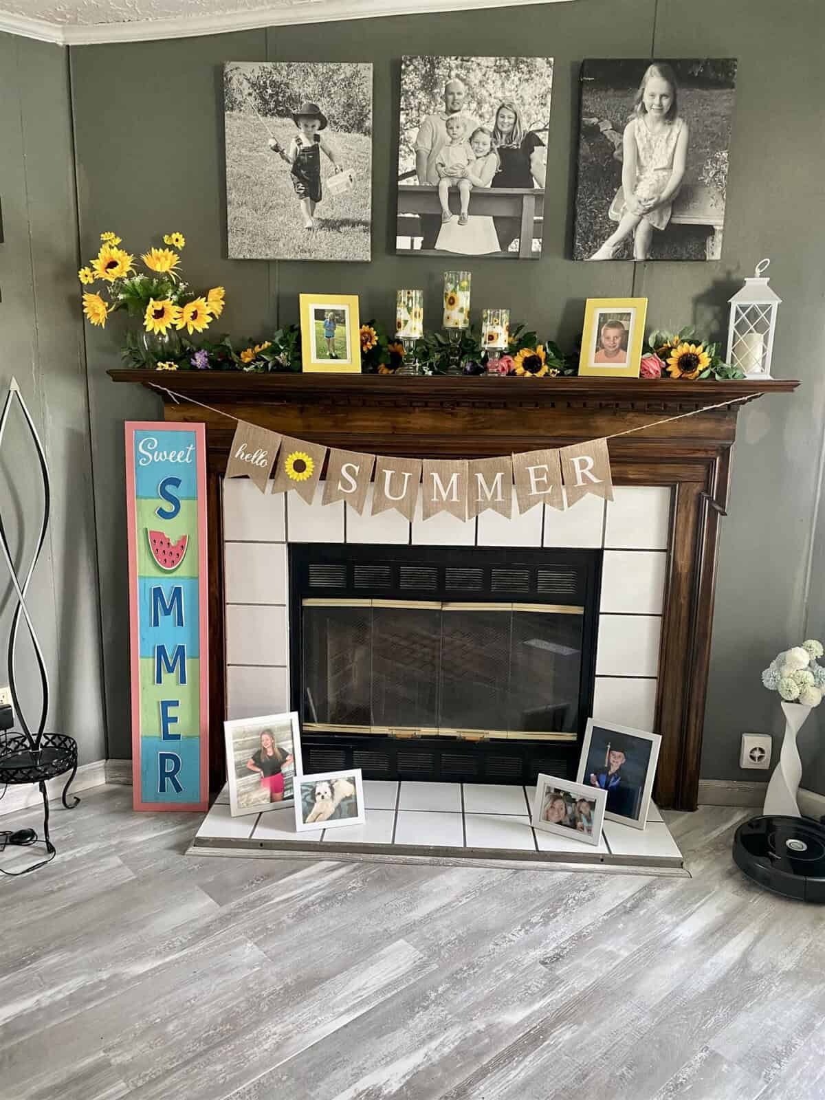 A fireplace is decorated with sunflower-themed decor and a "Hello Summer" banner. Family photos in frames are arranged on the mantel and around the hearth. A vertical "Sweet Summer" sign leans against the wall. The decor creates a warm, seasonal atmosphere.