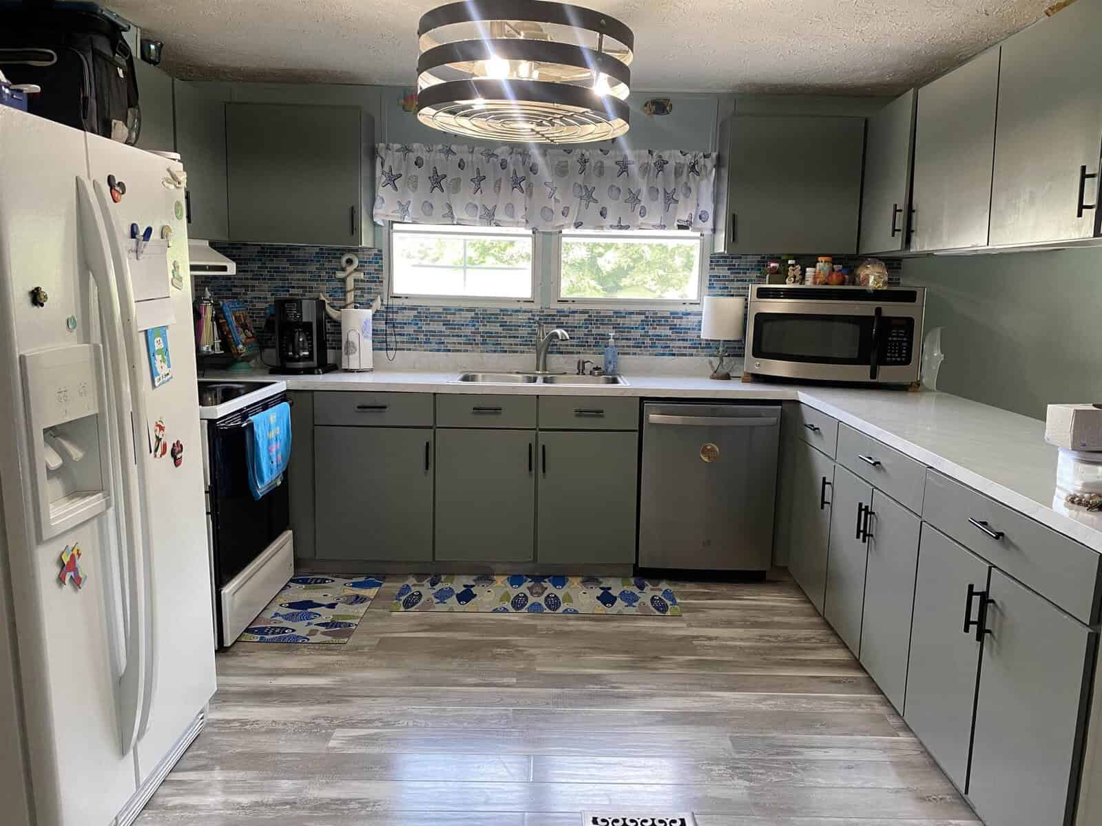 A modern kitchen with gray cabinets, white countertops, a stainless steel refrigerator, an oven, and a microwave. Above the sink, there are blue-patterned curtains and a window. The backsplash features a blue mosaic tile design. A circular light fixture hangs from the ceiling.