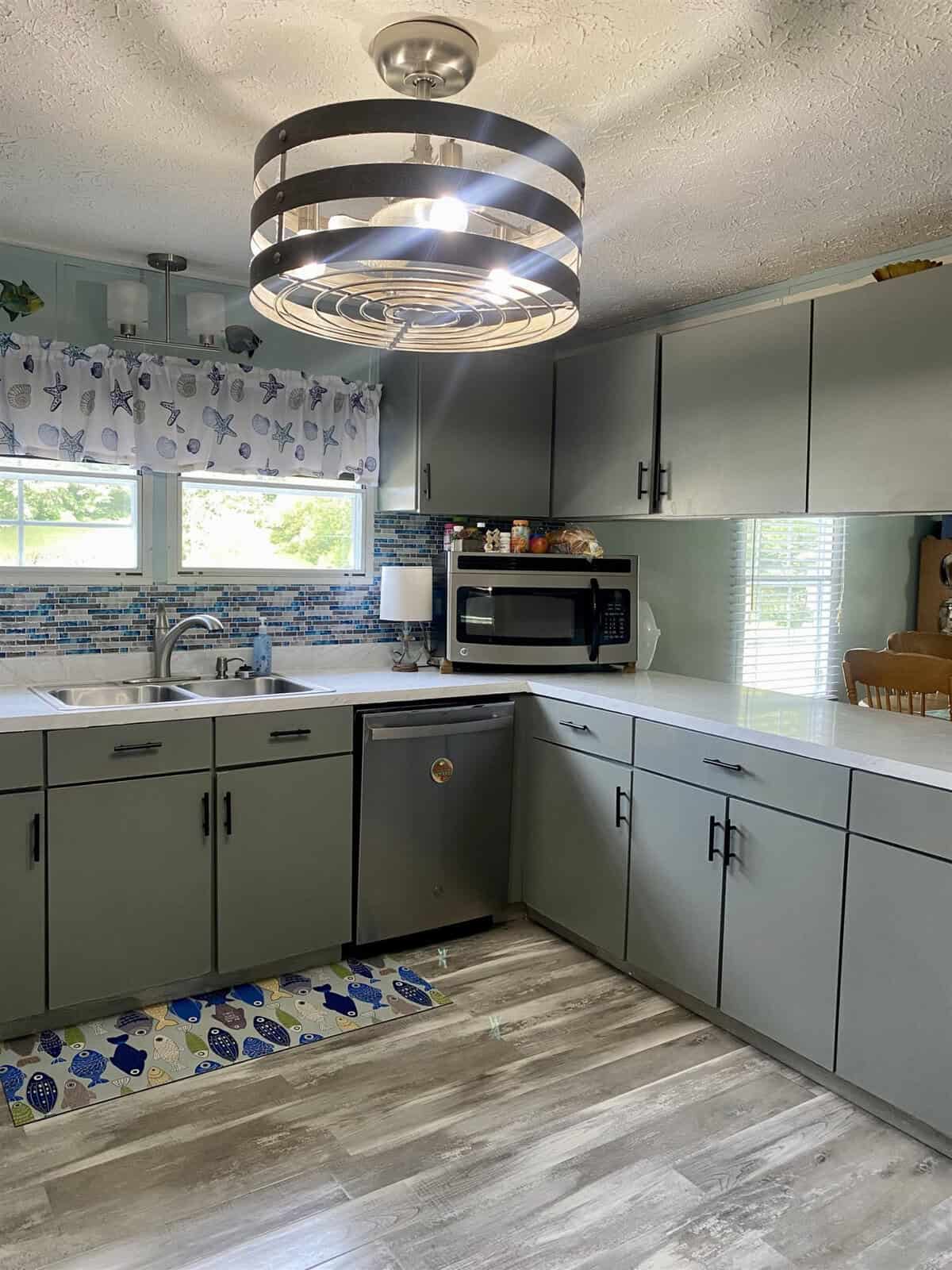 A modern kitchen with gray cabinets, light wood flooring, and a large circular light fixture. There is a backsplash with ocean-themed tiles, a window with patterned curtains, a dishwasher, and a microwave on the counter. The room is well-lit with natural light.