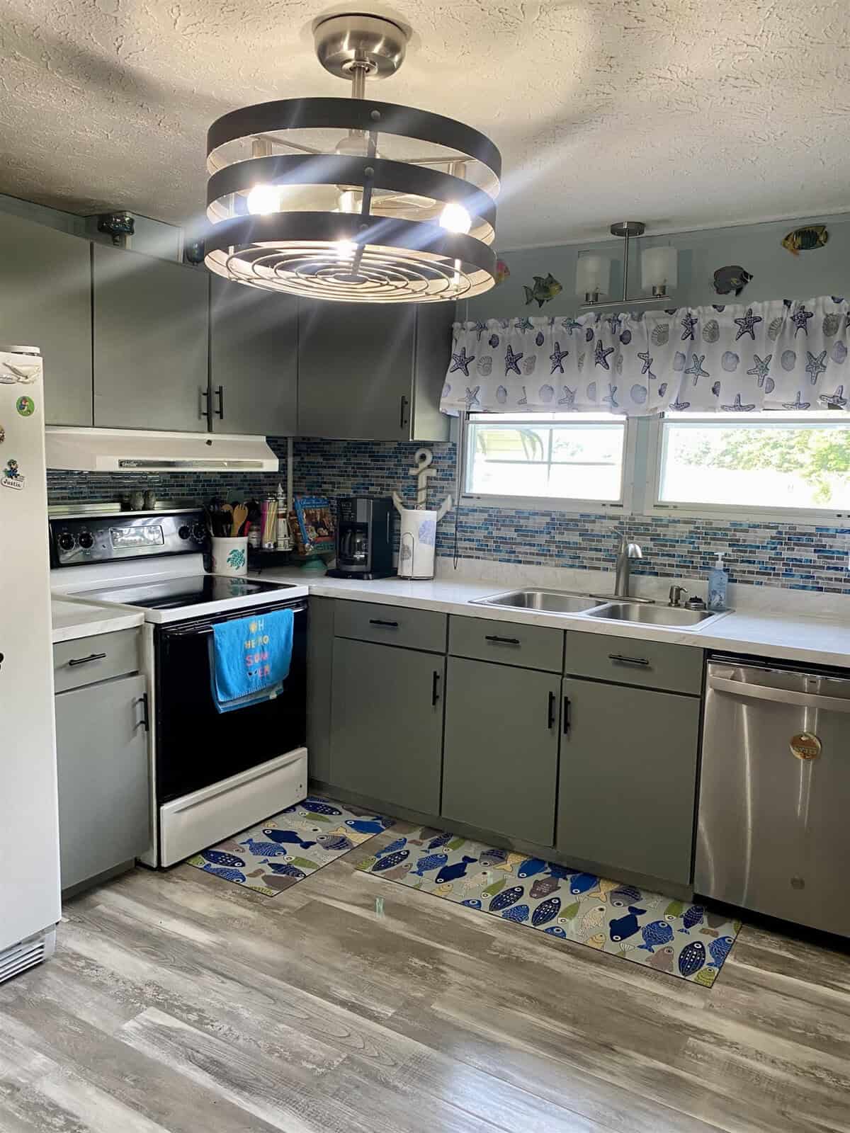 A kitchen with gray cabinets and wood-like flooring. The counter holds a toaster, coffee maker, and knife block. Above the stove is a modern light fixture. The window has a curtain with a nautical theme, and the blue backsplash complements the decor.