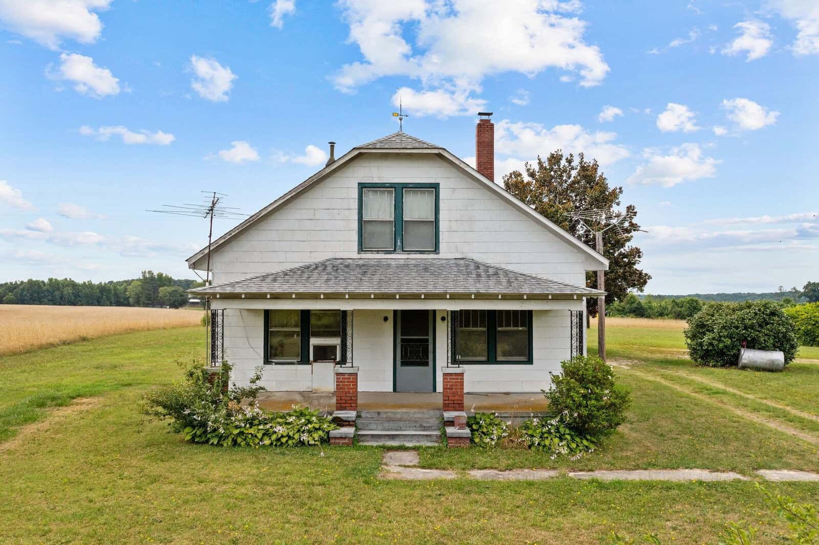 A two-story white house with a front porch, nestled in a rural setting with a clear sky, and surrounded by open fields.