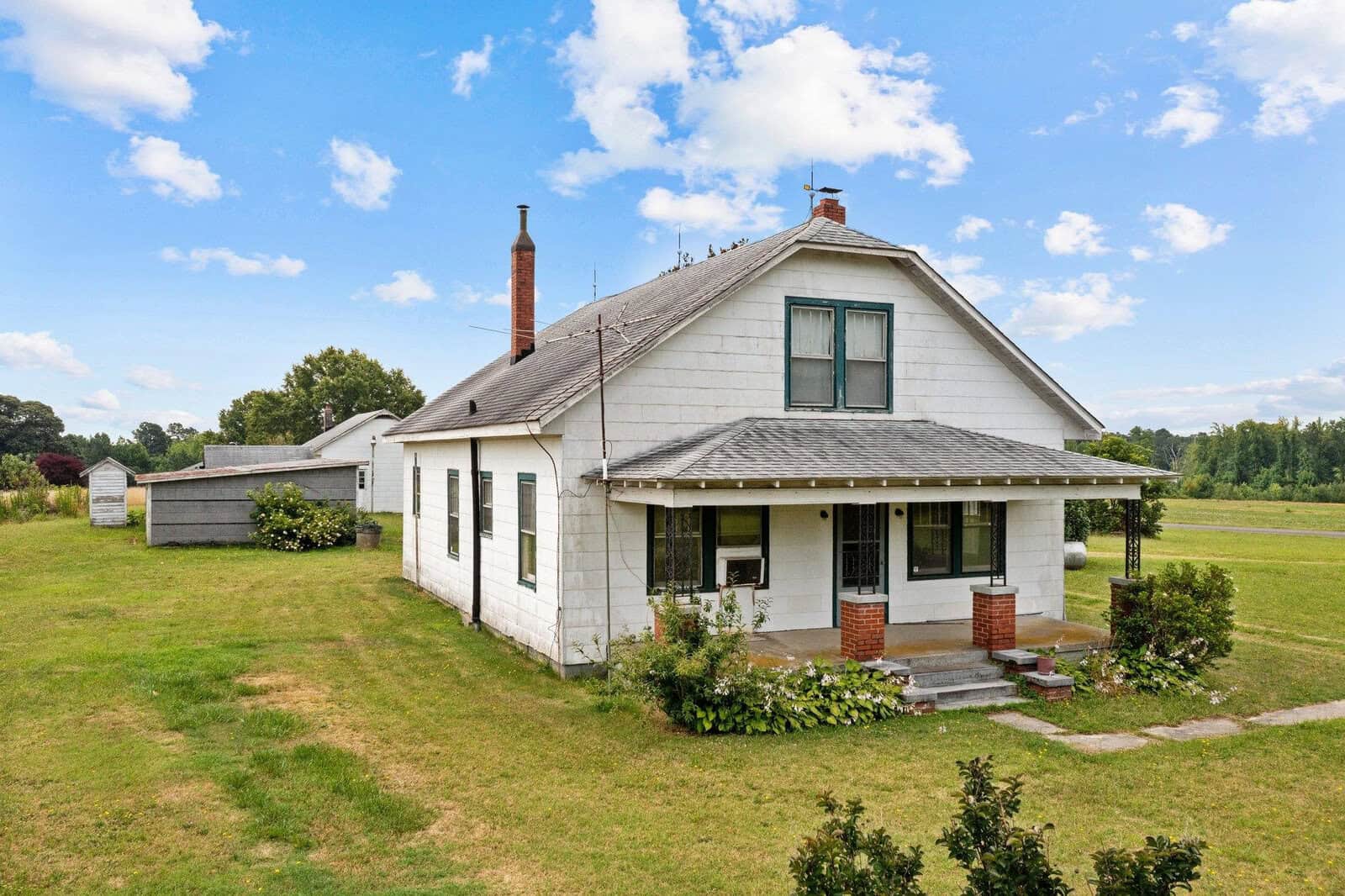 A white, two-story farmhouse with a gabled roof and a brick chimney, featuring a covered front porch, surrounded by a grassy lawn with a clear sky above and an outbuilding in the background.