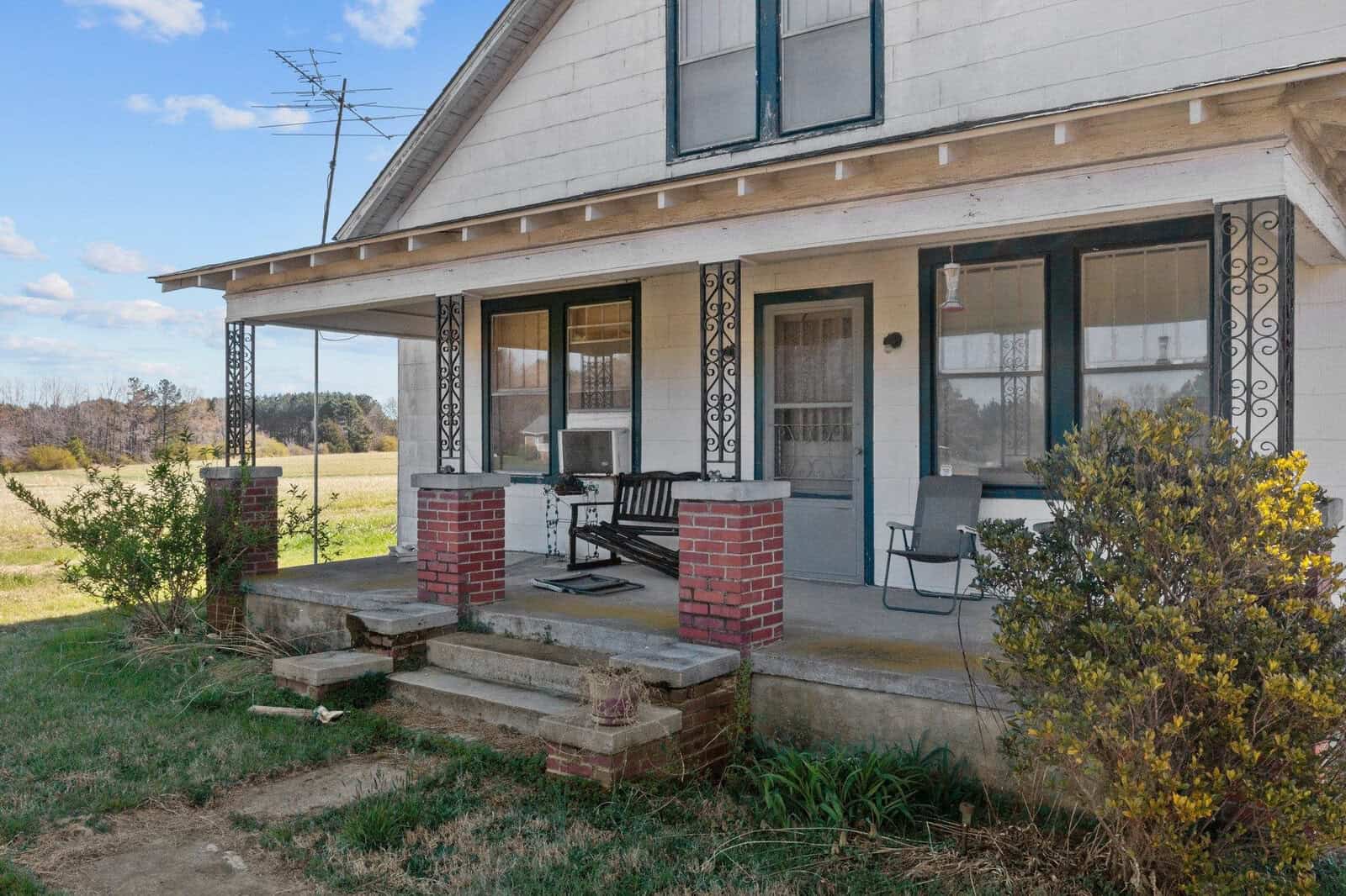 A rural two-story house with a brick foundation, featuring a covered front porch with a bench and a metal chair. the property is surrounded by open fields and there is a clear sky above.