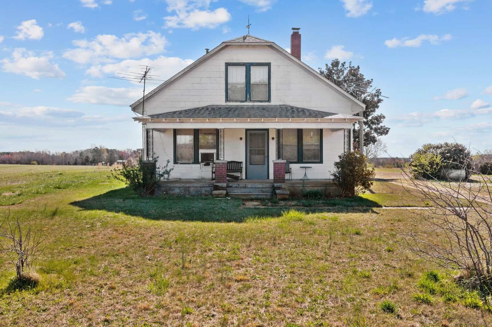 A two-story white house with a gabled roof and a front porch, set on a grassy plot with a clear sky above and open land in the background.