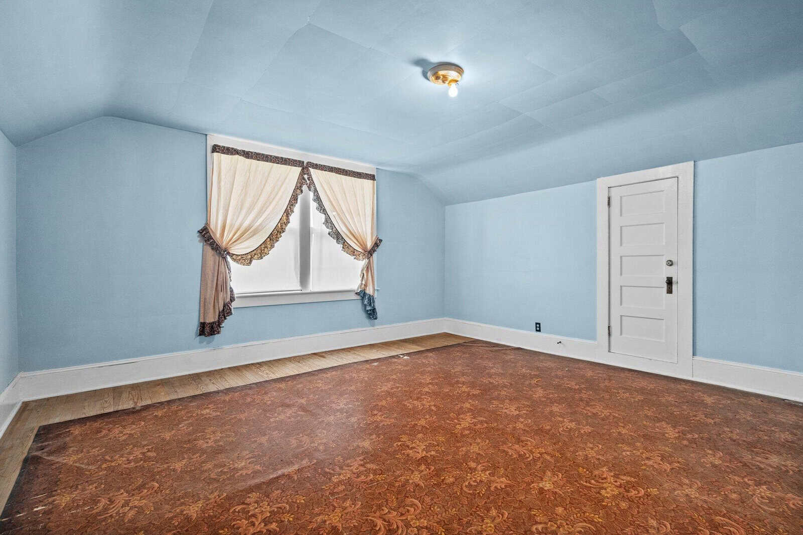 Empty room with blue walls, a patterned brown floor, a single window with draped curtains, and a closed white door.