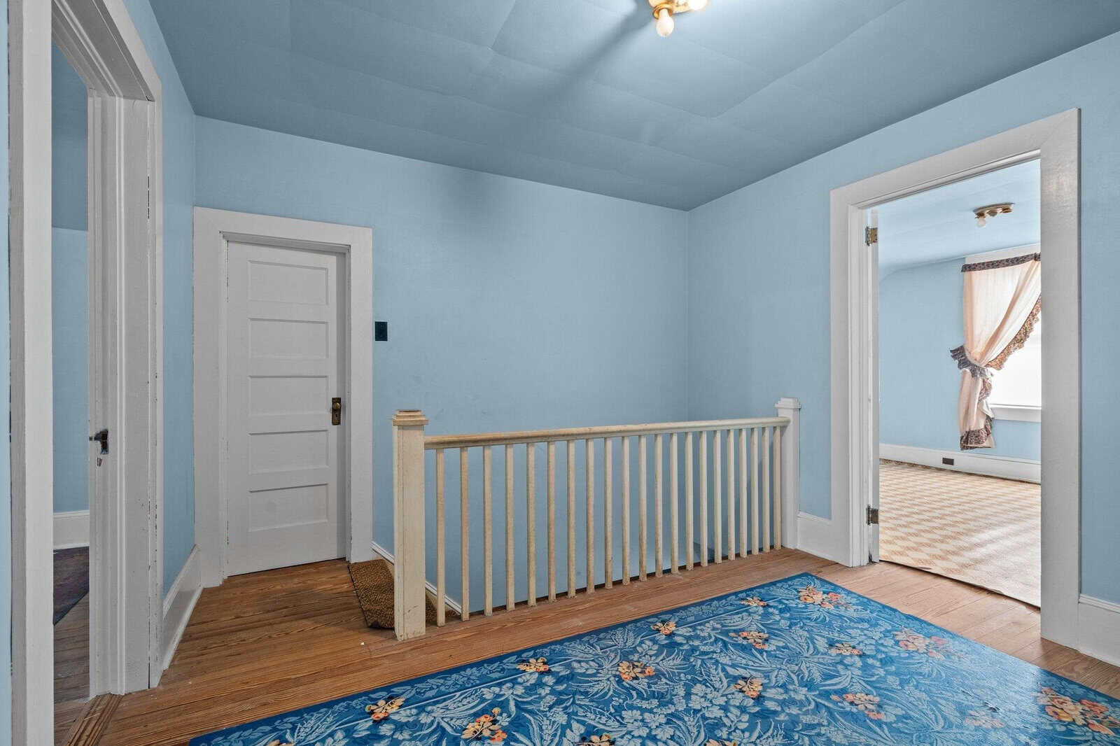 Empty hallway with light blue walls and a floral-patterned blue rug, featuring an open doorway to an adjoining room and a closed white door.