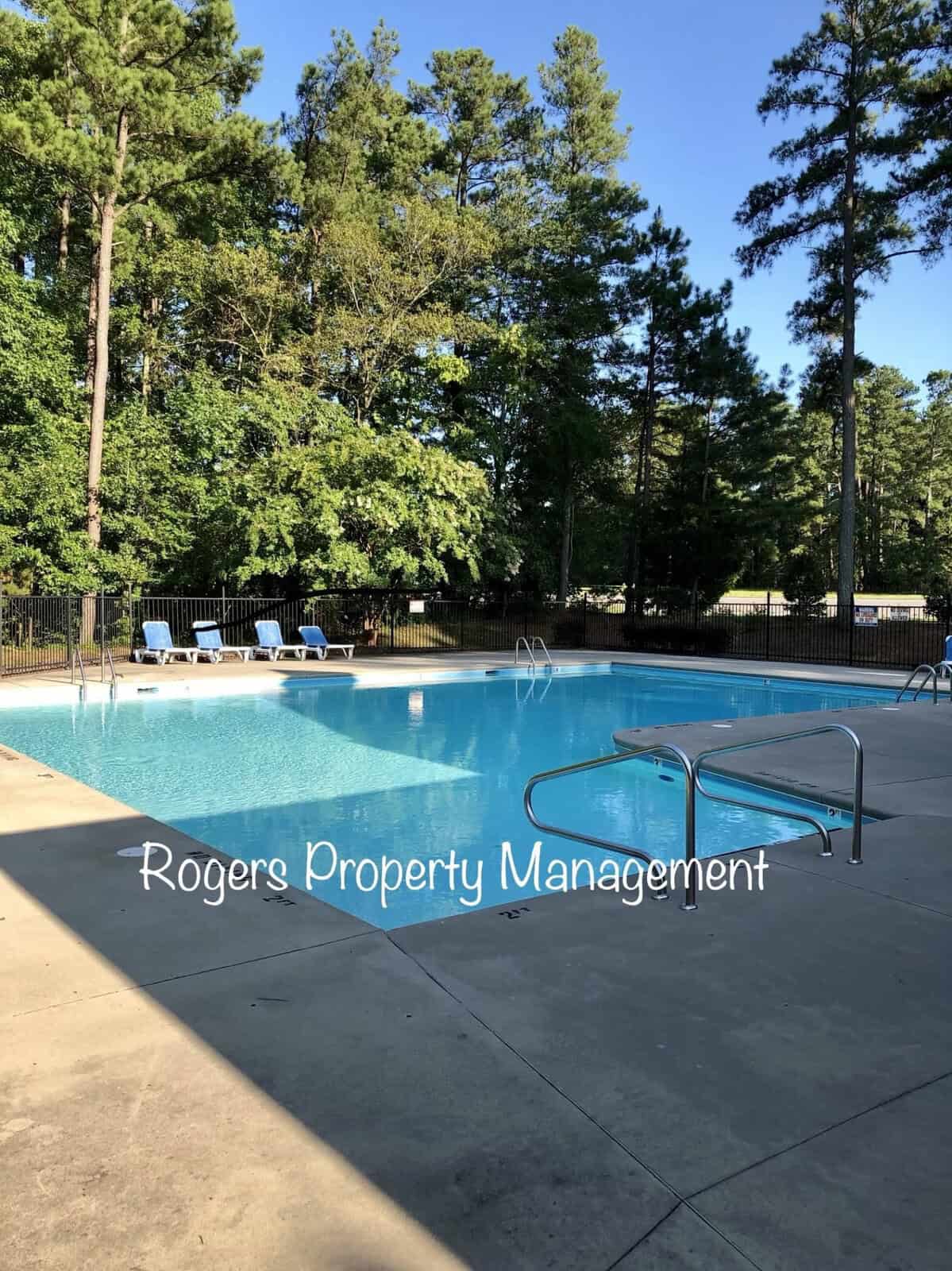 An outdoor swimming pool surrounded by trees with lounge chairs and stainless steel ladders, with a watermark reading "rogers property management.