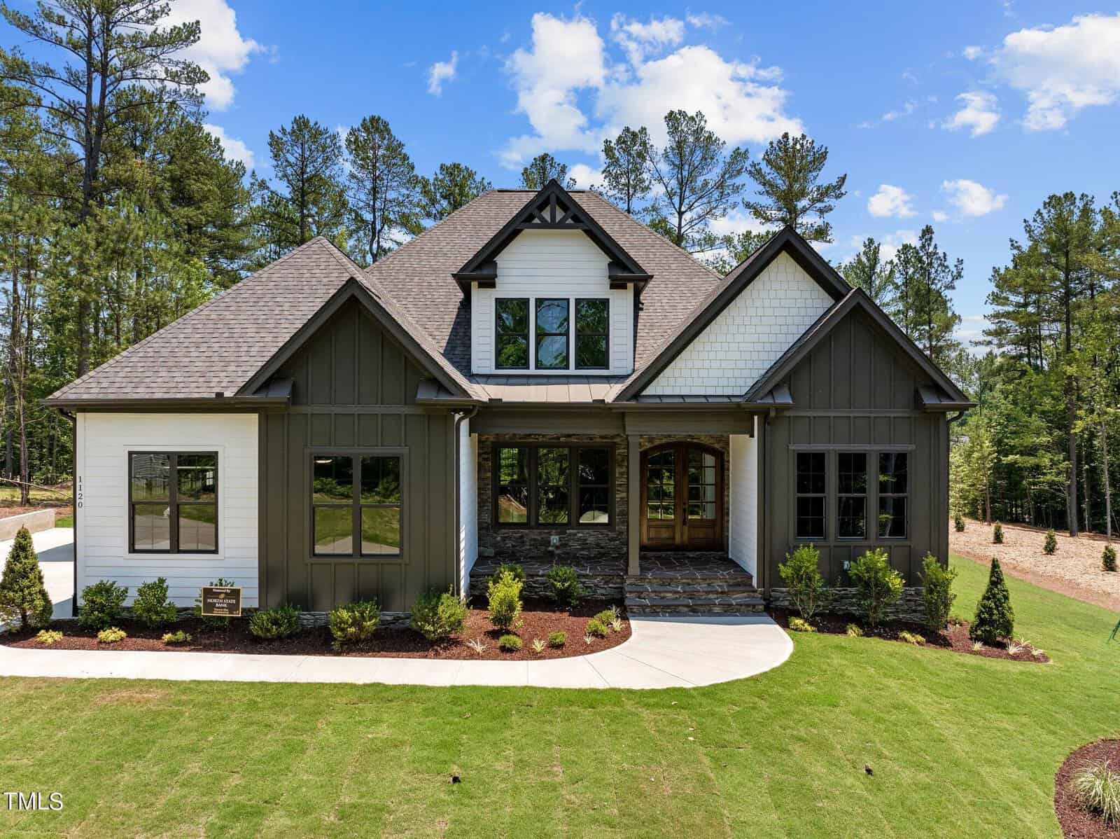 Nestled at 1120 Springdale Drive in Wake Forest, this modern suburban house boasts a gabled roof and striking white and dark green siding. The large front porch with stone steps opens to a neatly landscaped lawn, all under the serene backdrop of a clear blue sky.