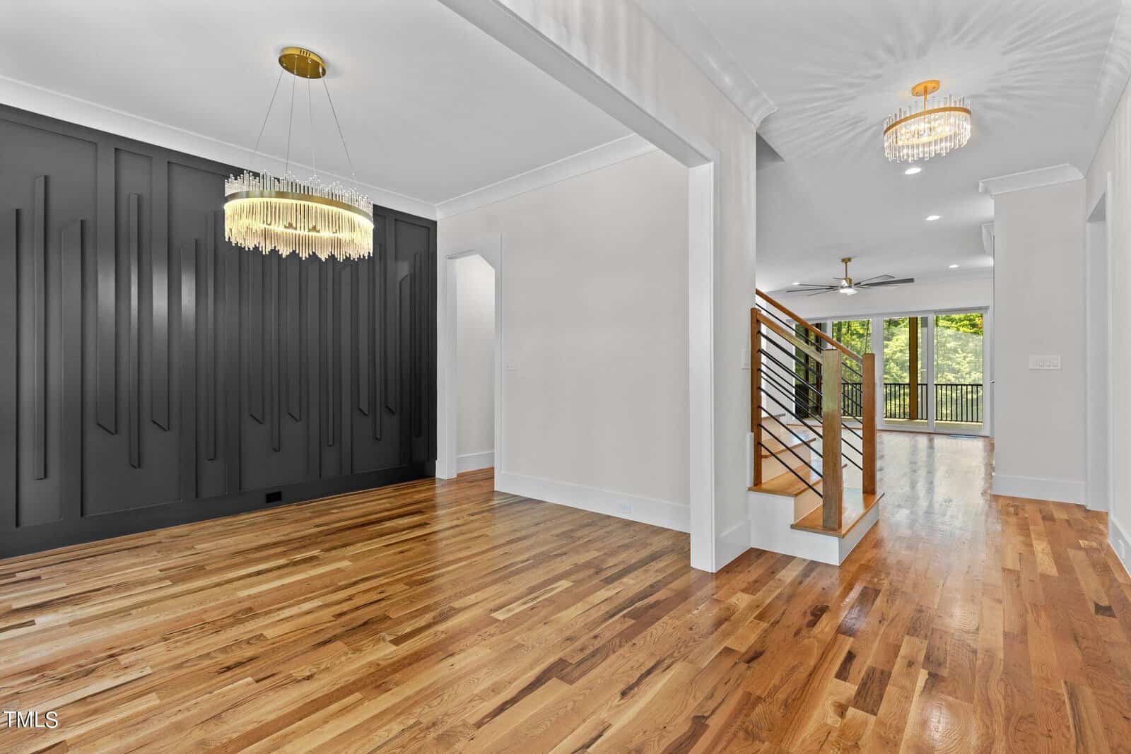 Interior view of a modern house with a dark accent wall and wooden flooring. A gold chandelier hangs from the ceiling. Staircase with metal railings leads to an open area with large windows and another chandelier.
