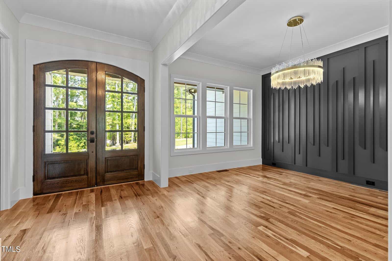 A bright room with large wooden double doors and a hardwood floor. To the right, there's a wall with vertical decorative panels and a hanging chandelier. Three windows on the back wall allow natural light to enter.