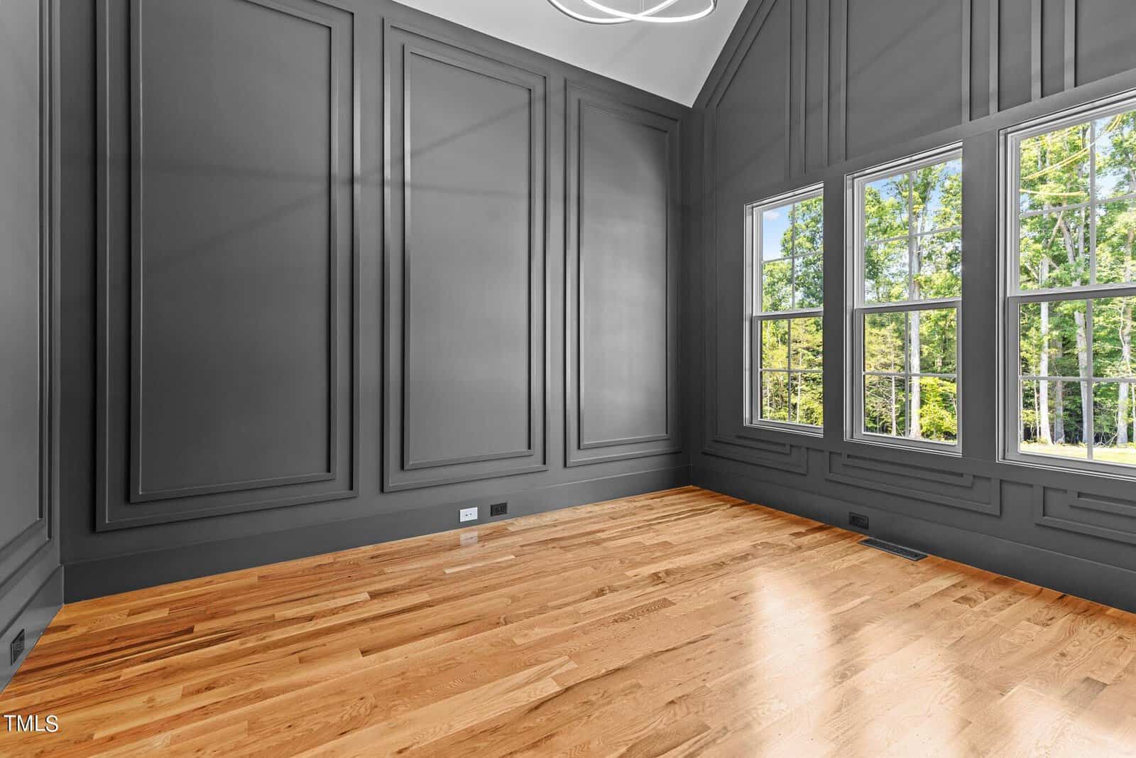 A spacious empty room with large windows letting in natural light. The walls are paneled in dark gray, and the floor is polished wood. The ceiling is vaulted, and a modern light fixture hangs from the center.