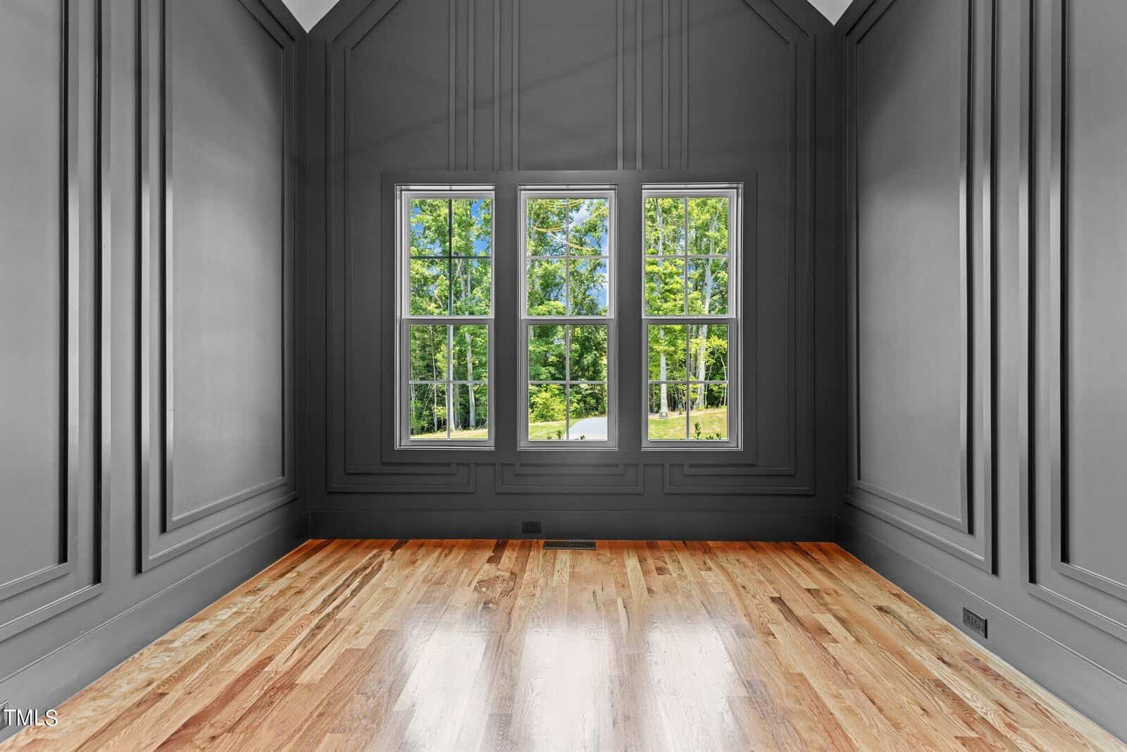 A bright, empty room with a high ceiling features gray paneled walls and a wooden floor. Three large windows offer a view of green trees outside, allowing natural light to fill the space.