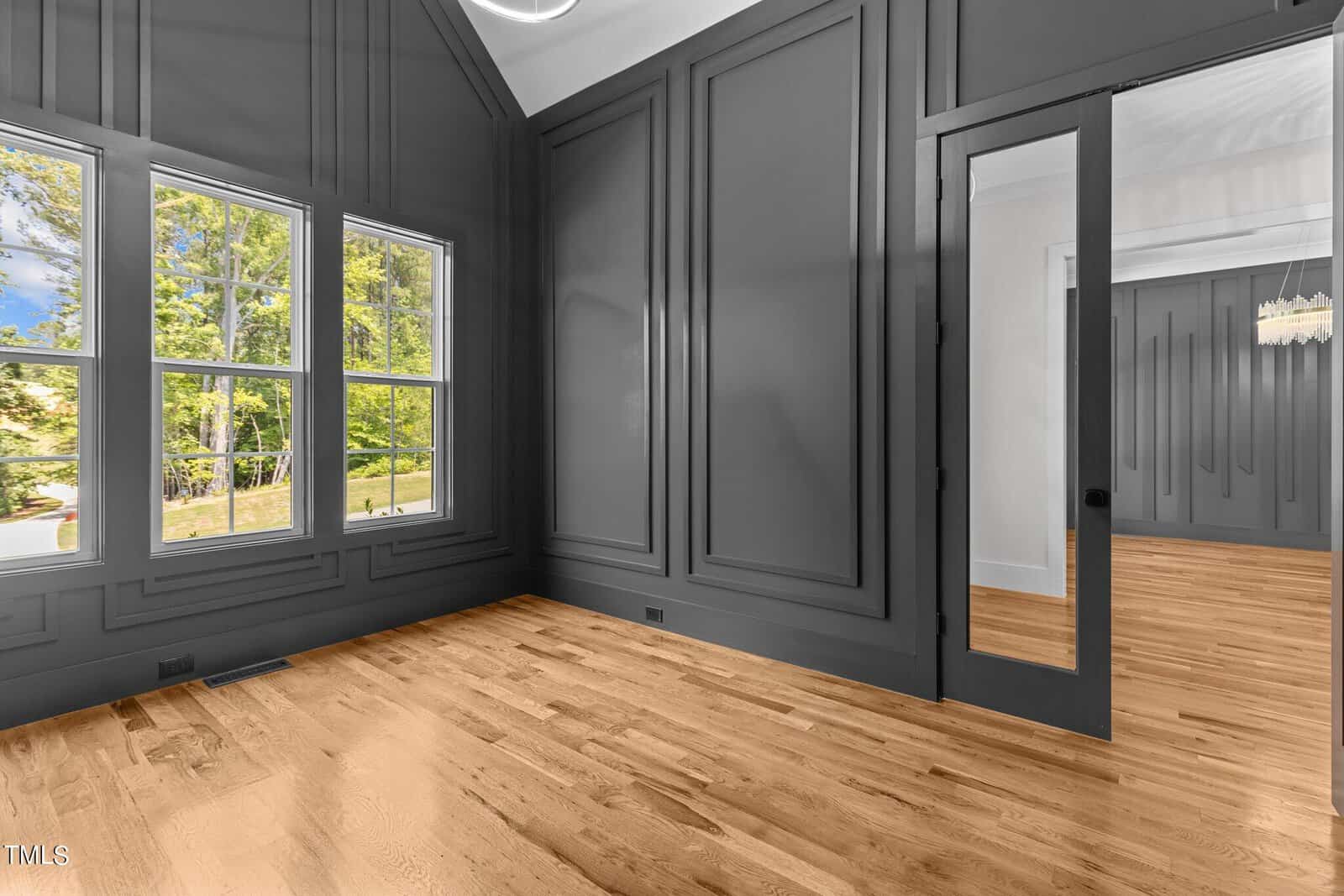 A room with gray paneled walls and a pitched ceiling. There's a large mirror on one wall and three windows on another, showing trees outside. The flooring is light wood, and a glimpse of an adjacent room with a chandelier is visible.