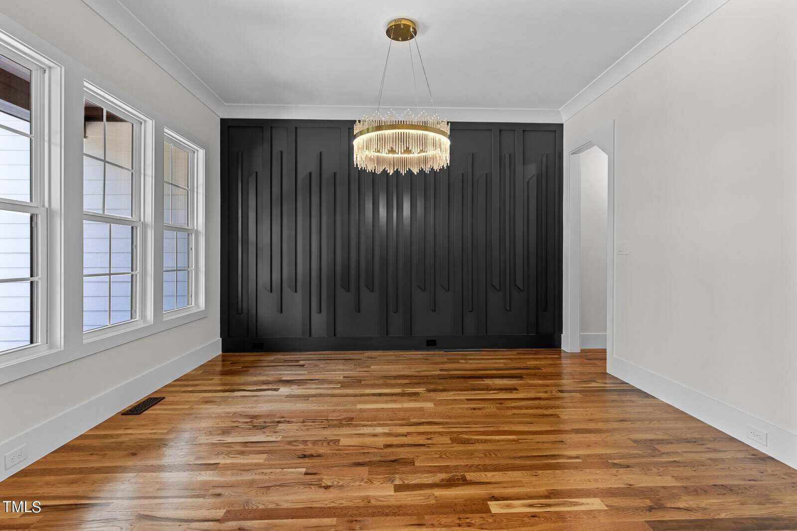 Empty modern room with dark wood accent wall, wooden floor, and large windows on the left. A distinctive chandelier hangs from the ceiling in the center. White walls contrast with the dark accent wall. An open doorway is visible on the right.