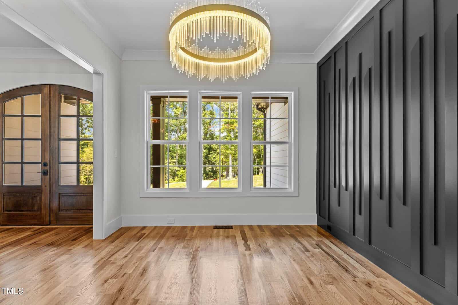 A room with light wooden flooring, a large window overlooking greenery, and a modern chandelier. The right wall features vertical black panels, while the left door is wooden with glass panels.