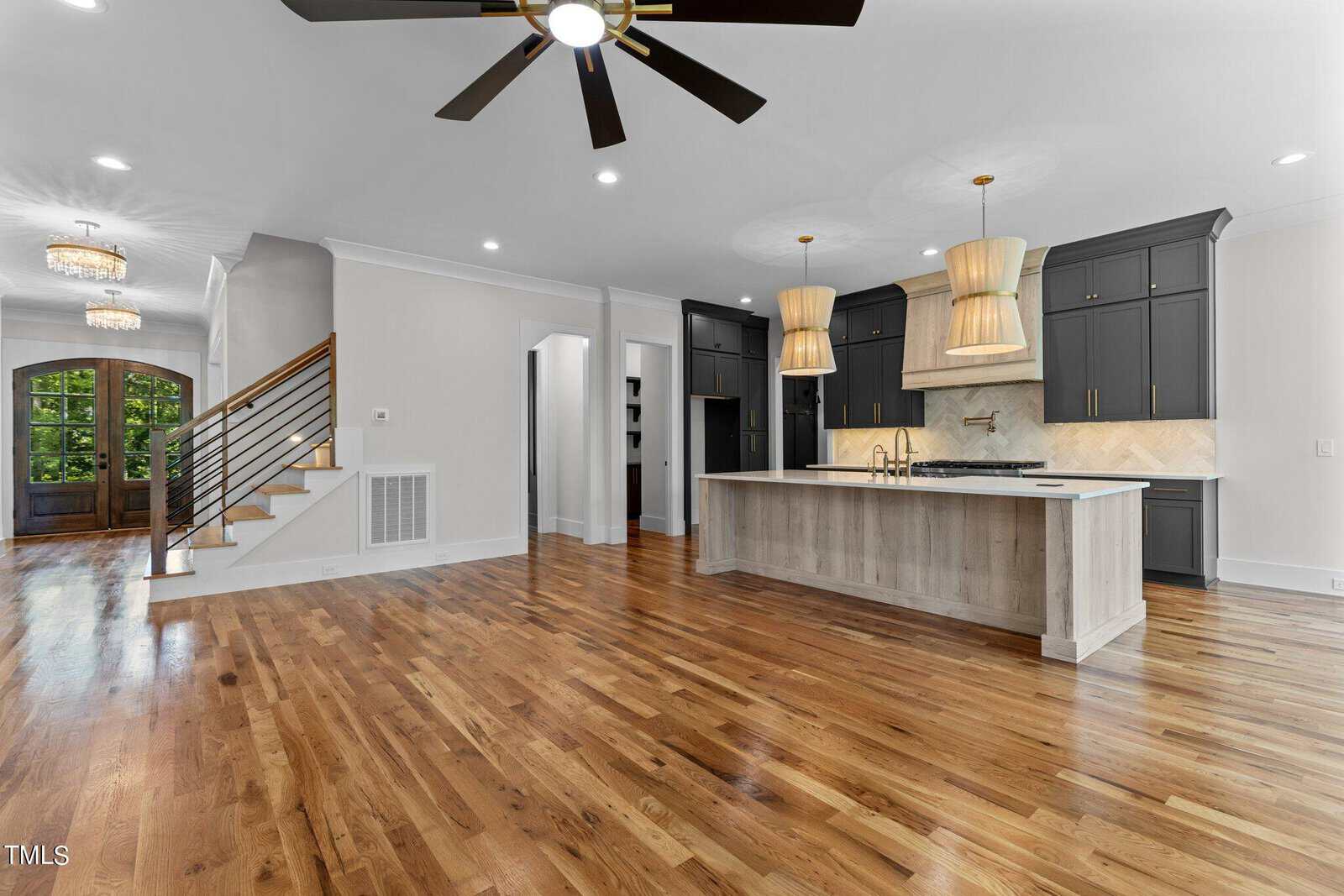 A spacious open-plan kitchen and living area with hardwood floors. The kitchen features a large island with a sink, modern appliances, dark cabinets, and hanging light fixtures. A staircase with a wooden railing leads to the upper level.