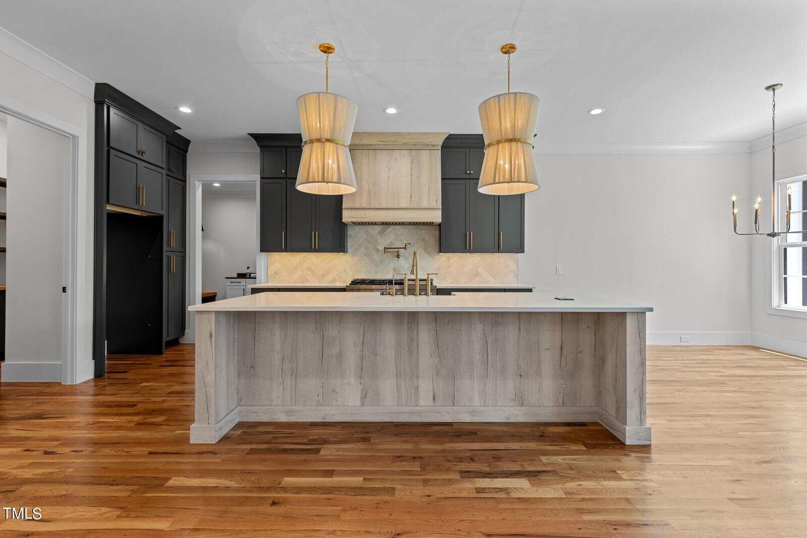 A modern kitchen with hardwood floors features a large island with a light wood finish and dual sinks. Two pendant lights hang overhead. The background has dark cabinetry and a light-colored backsplash. A chandelier is visible in the adjacent dining area.