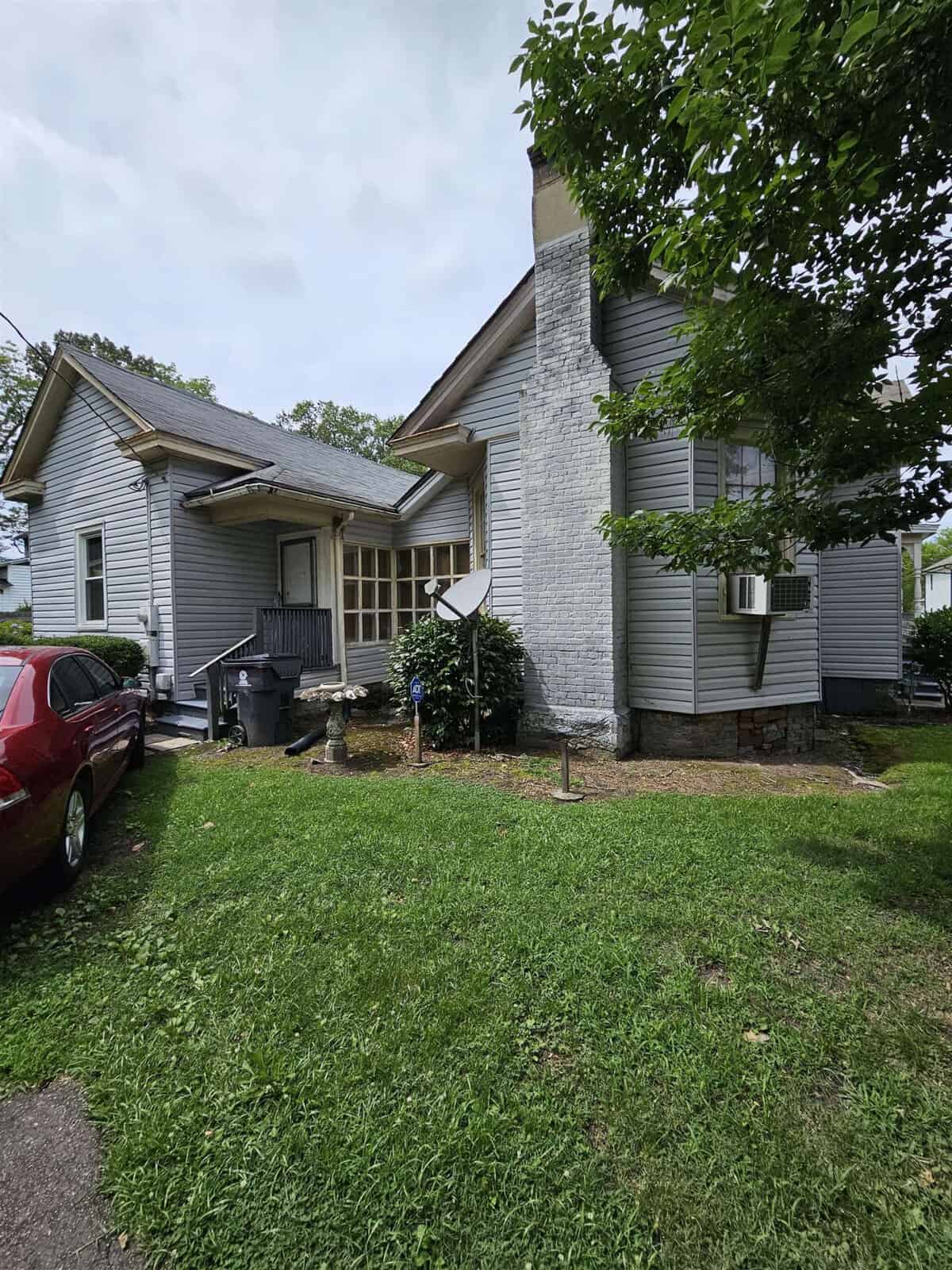 A two-story house with gray siding and white trim, featuring a chimney and a front porch, with a red car parked on the left side. trees and a lawn are visible in the foreground.