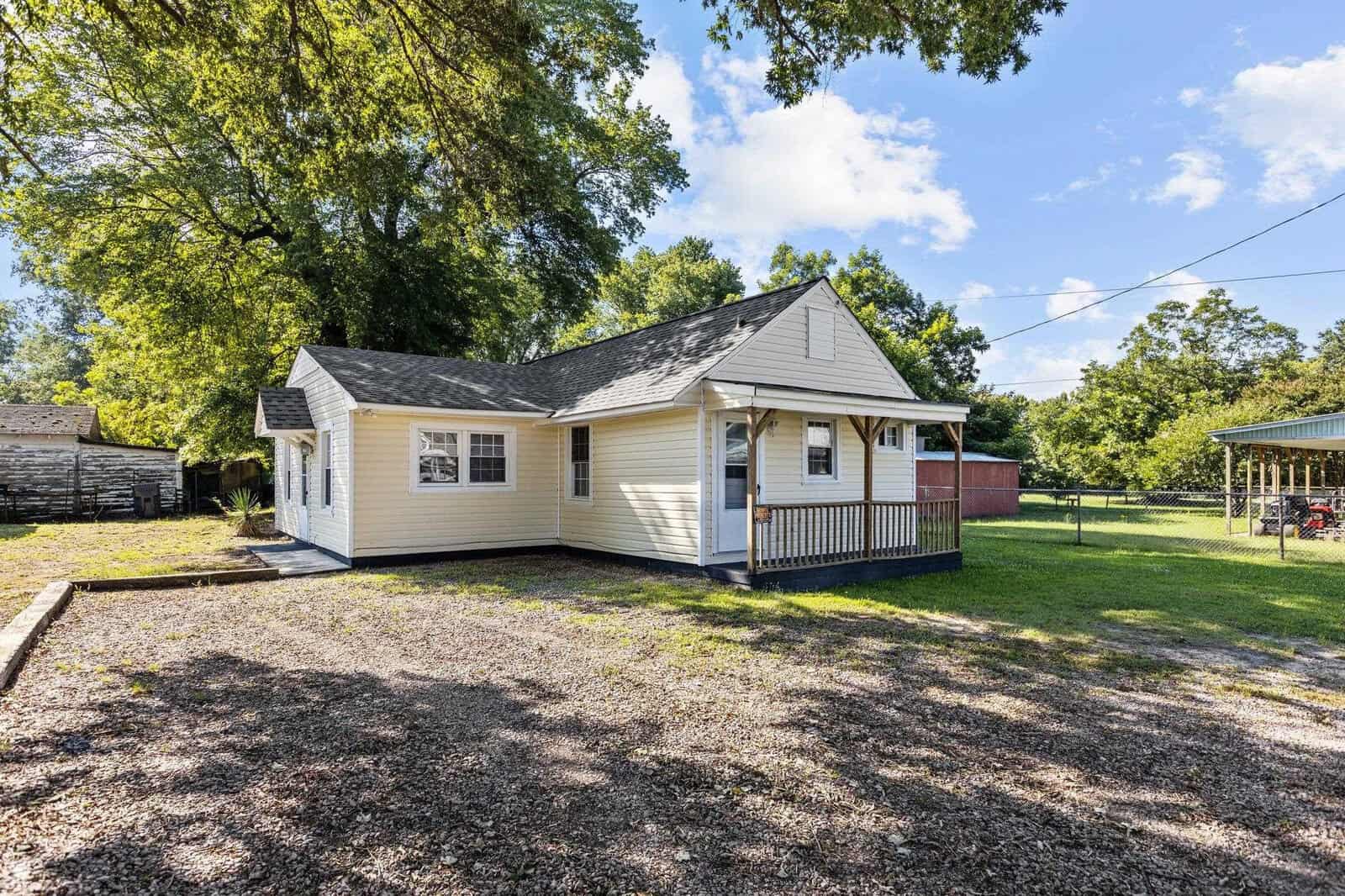 A small, single-story house with beige vinyl siding and a dark shingled roof, featuring a front porch with white railings. the property includes a gravel front yard, trees in the background, and a clear sky above.