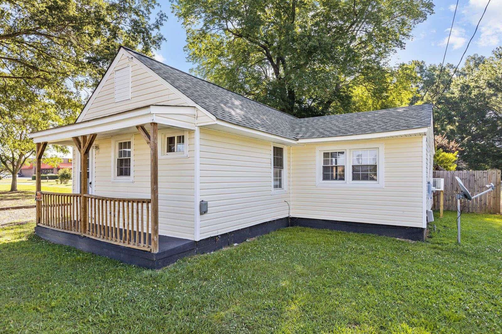 A small, white single-story house with a wooden front deck and a sloped roof, surrounded by a grassy yard with a clear sky above.