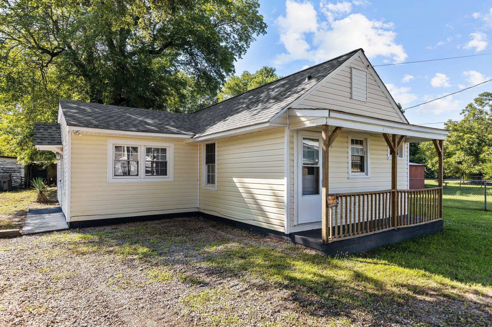 A small, single-story suburban home with a gabled roof, freshly painted beige siding, white trim, and a newly constructed front porch with a wooden railing. the house is surrounded by a modest yard with a gravel driveway on the left side.