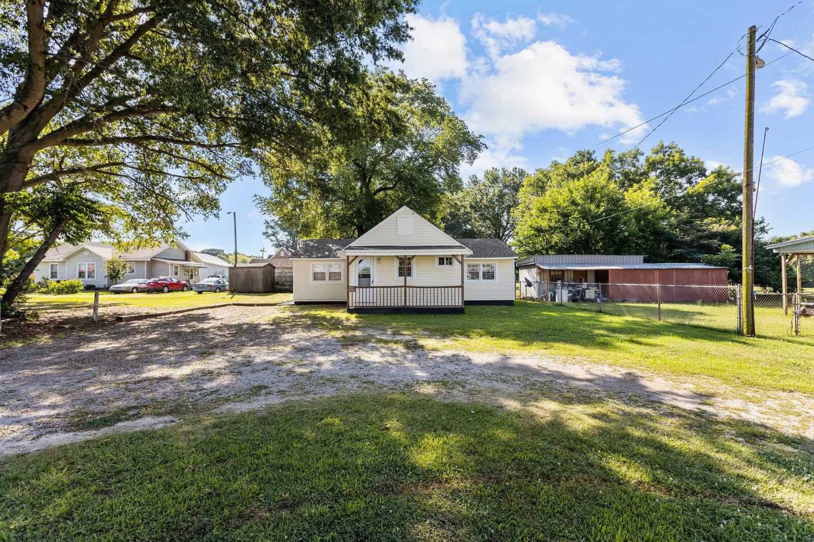 A small, single-story yellow house with a white trim is situated in a spacious yard with scattered green patches of grass and a dirt driveway. surrounding the property are trees, a wooden fenced area, and neighboring houses under a clear sky.