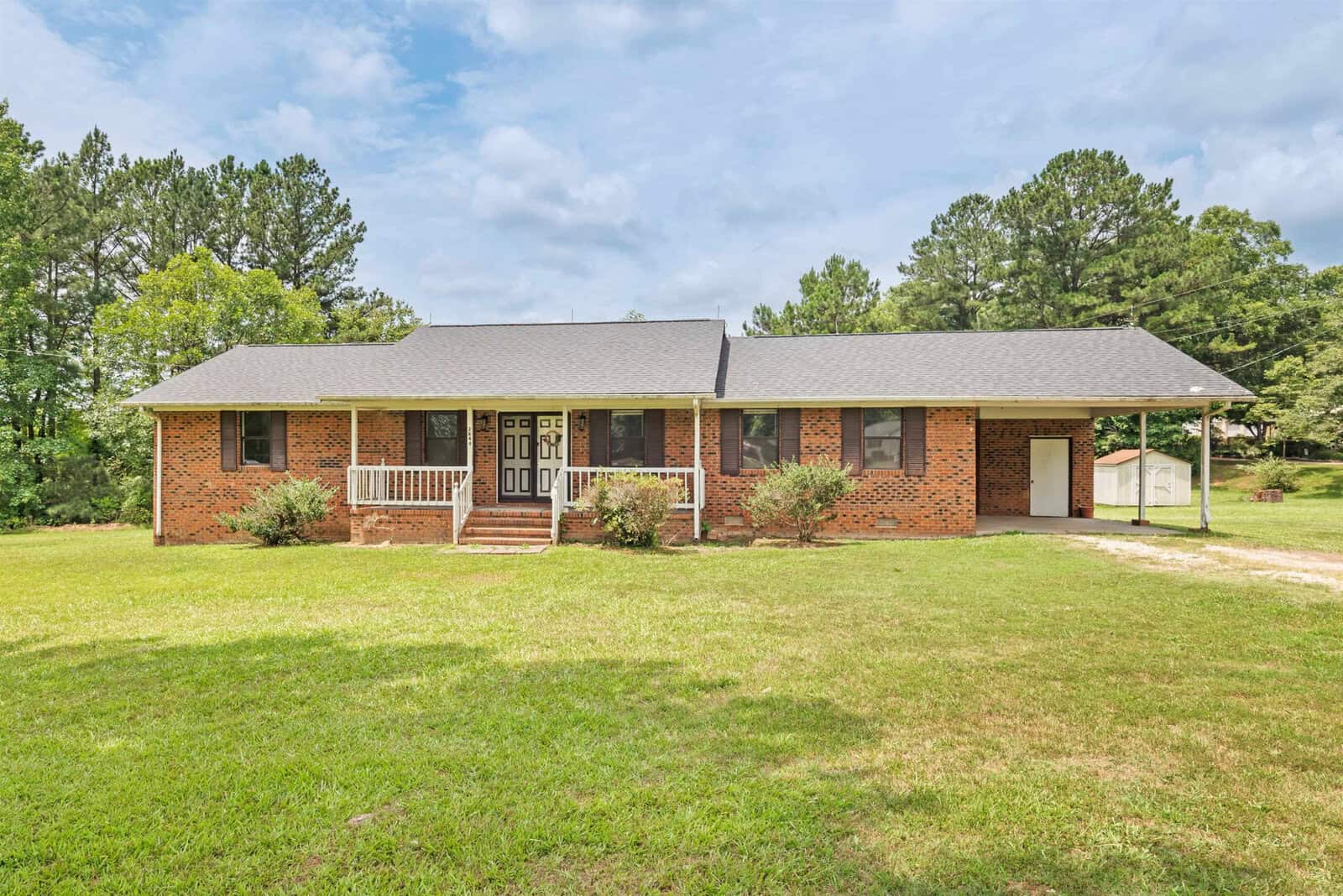 A single-story brick house with a dark gray roof and a front porch featuring white railings and steps. The house is surrounded by a grassy lawn with trees in the background. A small outbuilding is visible to the right. The sky is partly cloudy.