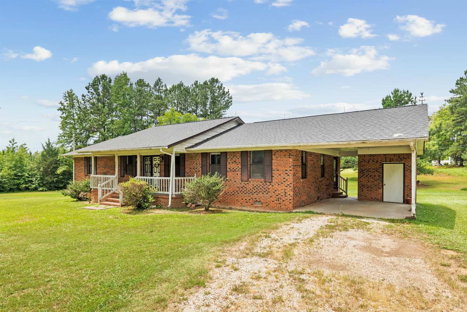 A single-story brick house with a covered front porch and white railings, surrounded by a grassy lawn. A carport is attached on the right side. Trees and a few bushes line the background on a clear, sunny day with scattered clouds.