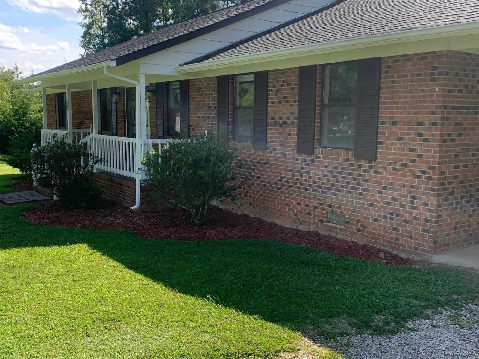 A single-story brick house with a front porch featuring white railing and brown shutters on the windows. The front yard is grassy with neatly trimmed bushes and a garden bed lined with red mulch along the foundation. Trees can be seen in the background.