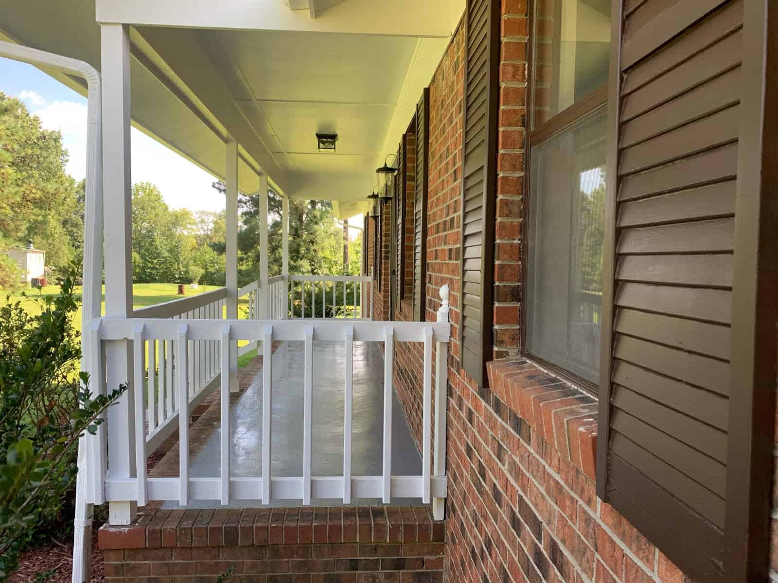 A porch with white railing attached to a brick house with brown shutters. The porch is surrounded by greenery with a lawn and trees visible in the background. The ceiling of the porch is white, and a light fixture is mounted near the entrance.