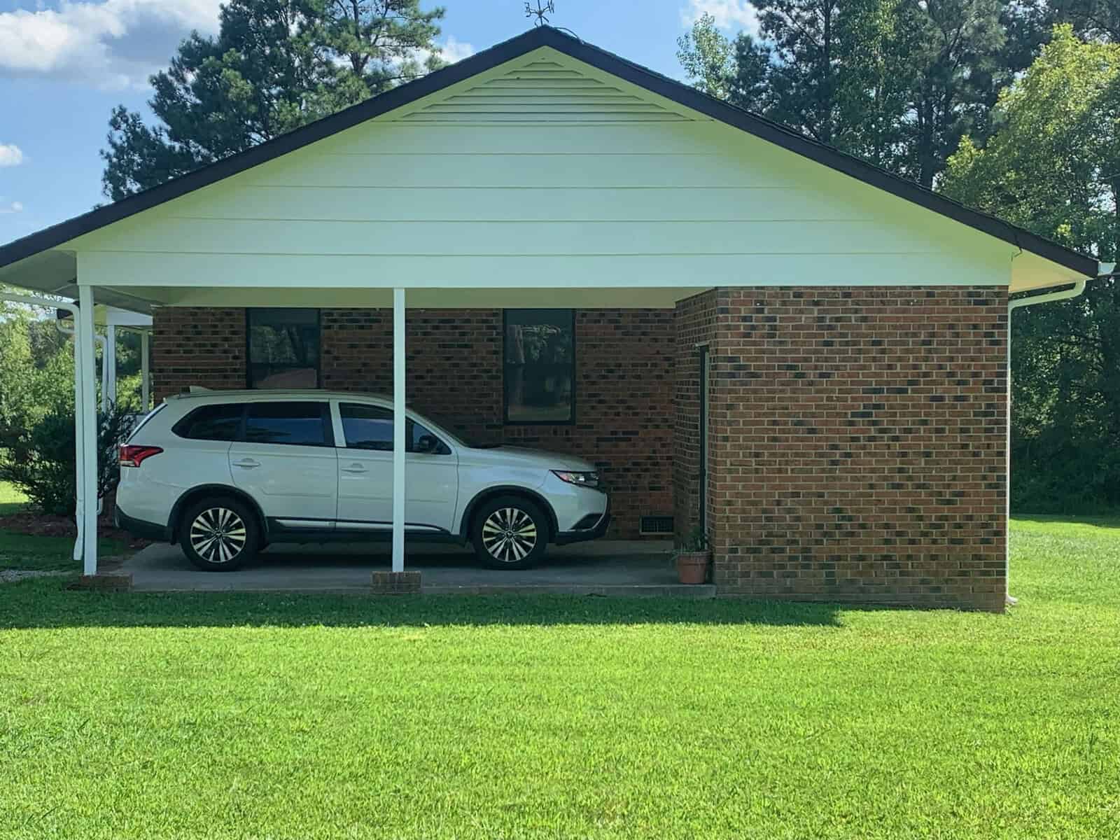 A white SUV is parked under an attached carport of a brick house. The house has a gabled roof with white siding at the top and red brick below. The lawn in front is well-kept and lush green, with trees visible in the background. The sky is partly cloudy.
