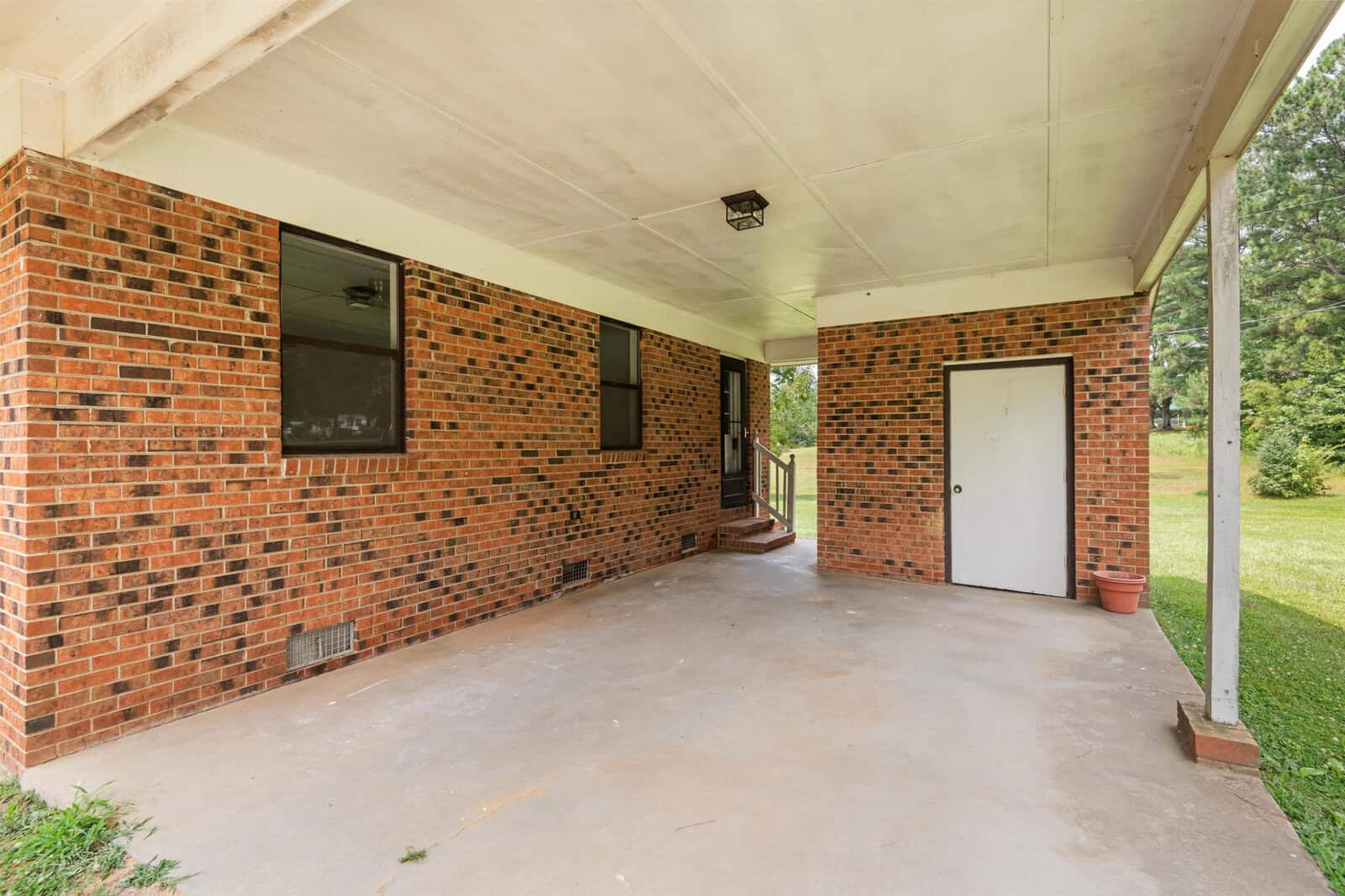 A brick house with a covered patio area. The patio has a concrete floor and a white ceiling. There are two windows, a door, and a small set of stairs leading to another door. A single potted plant is placed in the corner. The background features green grass and trees.