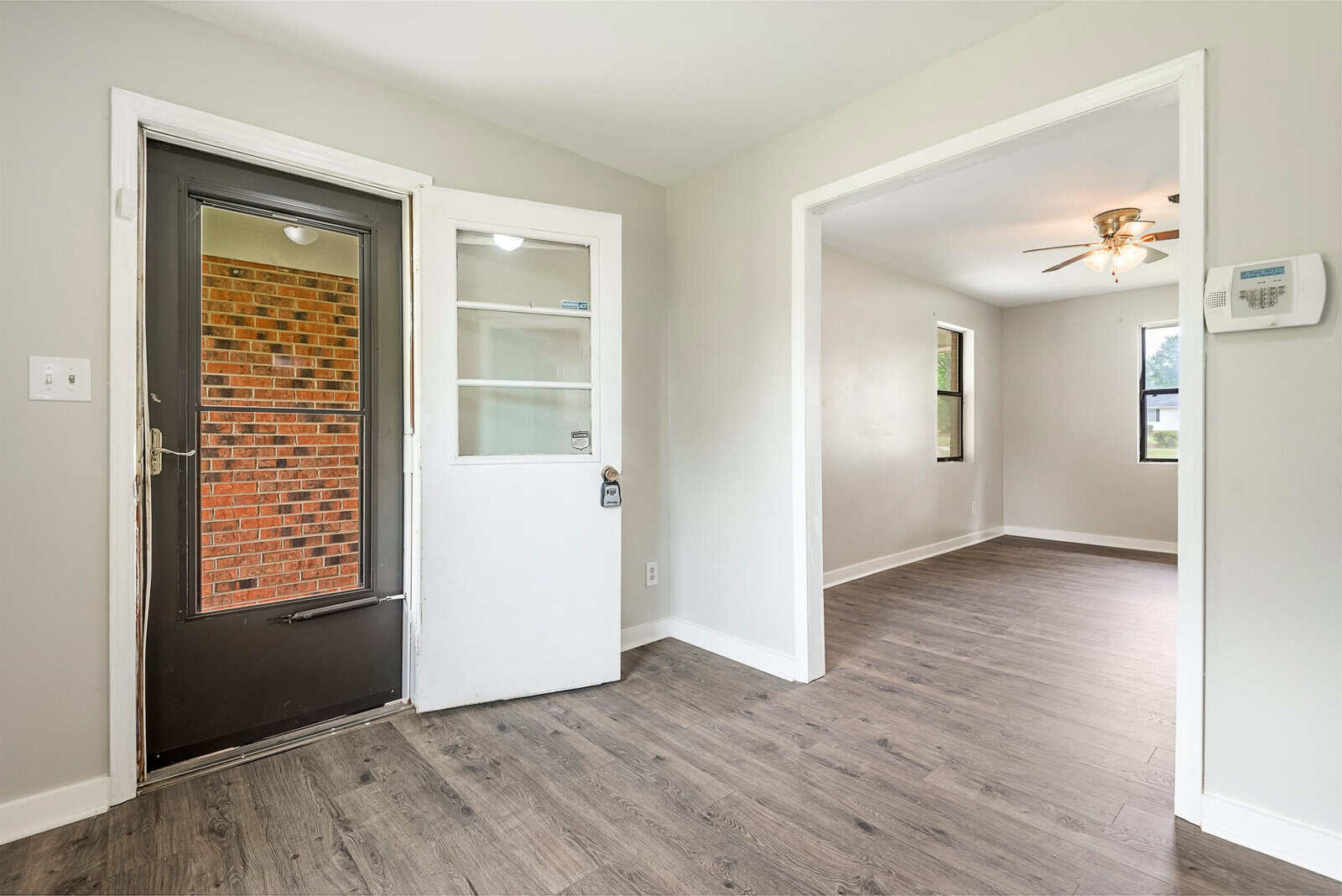 A small entryway with a black front door featuring a large window. Beyond it, there is a short hallway with light gray walls and dark wood flooring leading into a bright living room with windows, white walls, a ceiling fan, and a thermostat on the wall.