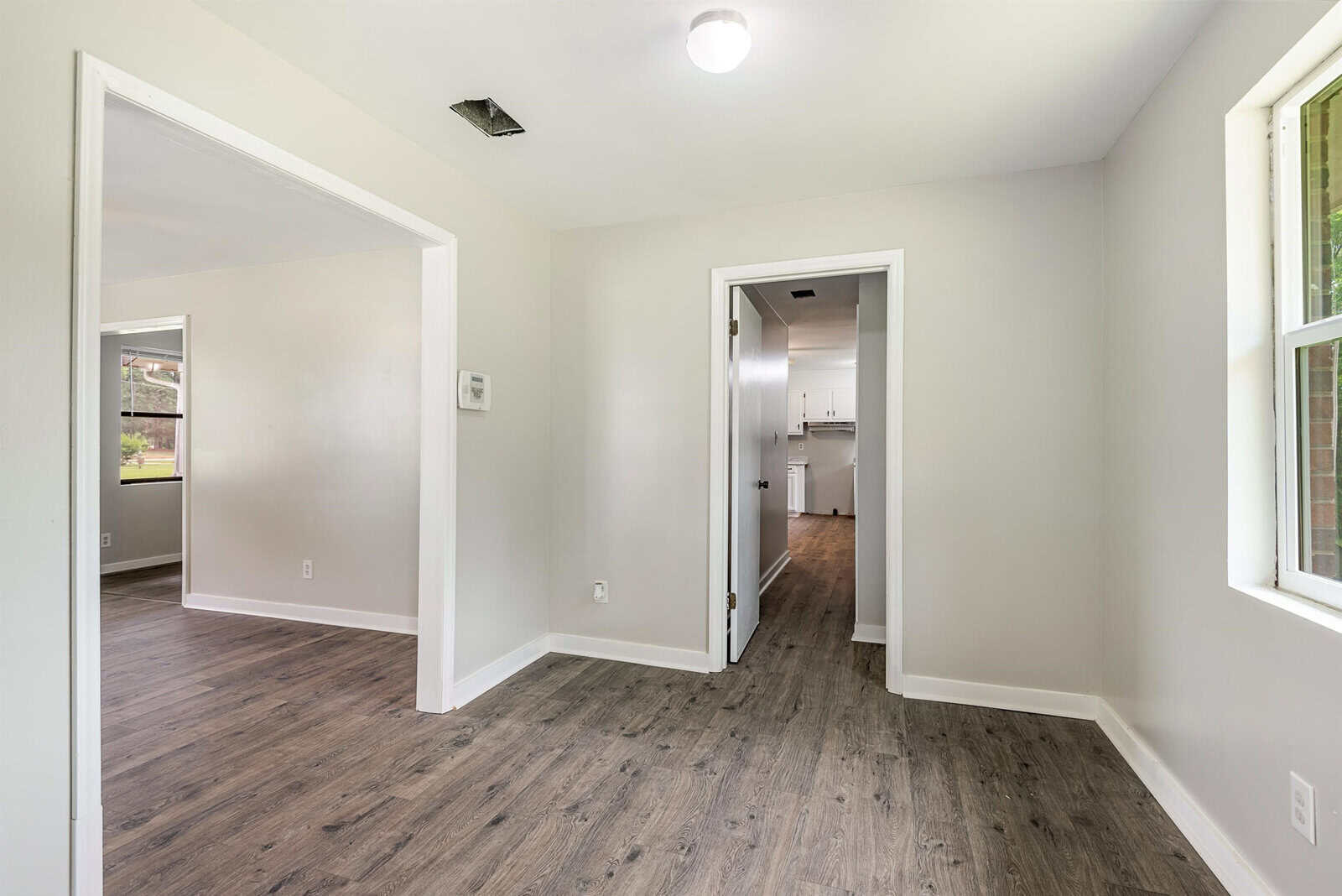 A bright, empty room with light gray walls and wooden flooring features two open doorways on the left, revealing adjacent rooms. A hallway leads to a white kitchen visible in the background. Large windows allow natural light to fill the space.