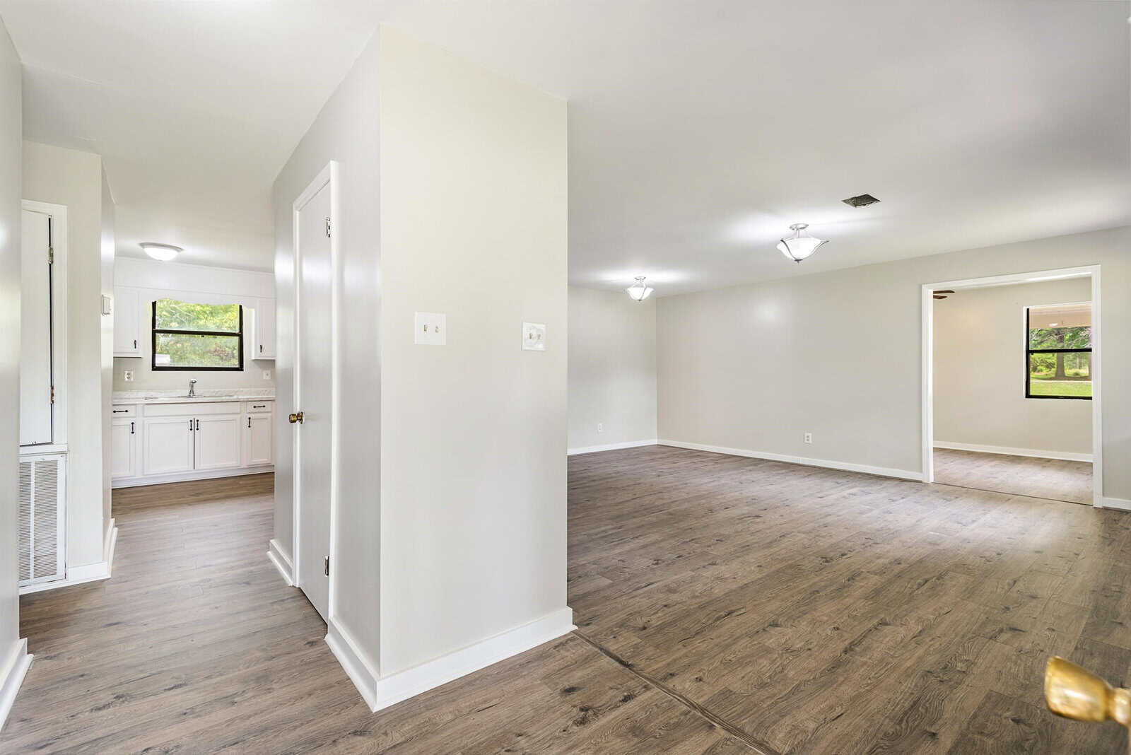 A modern, empty interior with light gray walls and wood laminate flooring. The view shows a kitchen to the left with white cabinets and a window, a spacious living area, and another room in the background with natural light from large windows.