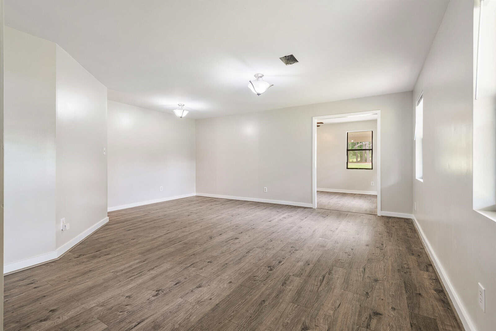 A spacious, empty room with light-colored walls and hardwood flooring. The room is well-lit with modern ceiling lights and features an open doorway leading to another room with a window that offers a view of greenery outside.