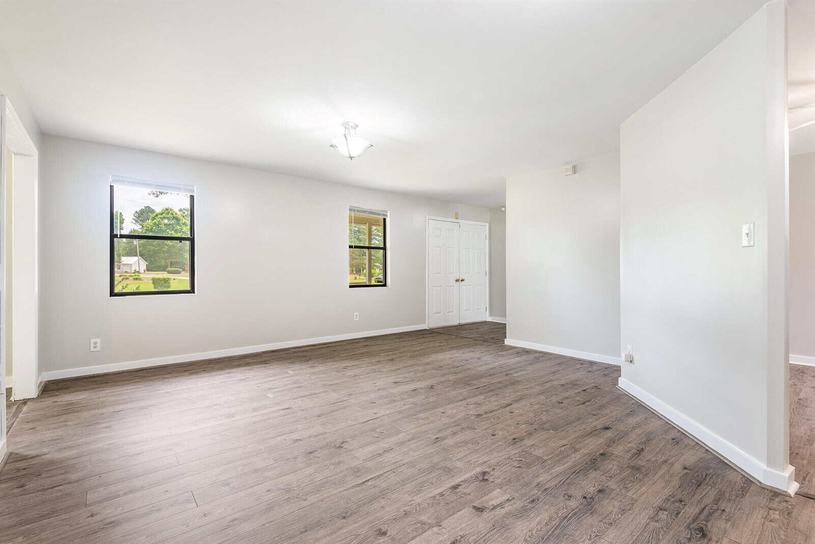 A modern, unfurnished living room with light gray walls, a large window letting in natural light, and dark wood flooring. The room is spacious, with an entryway and a white double door closet visible. The ceiling features a small, central light fixture.