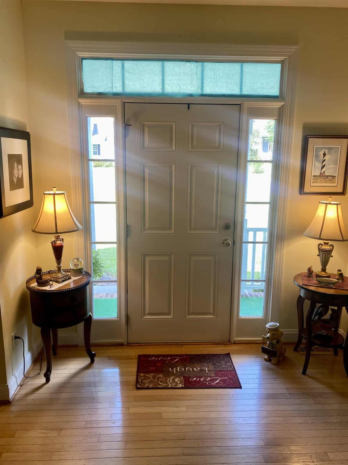 A warmly lit foyer featuring a white front door with a transom window, flanked by decorative table lamps on small tables, and a red welcome mat on the wooden floor.