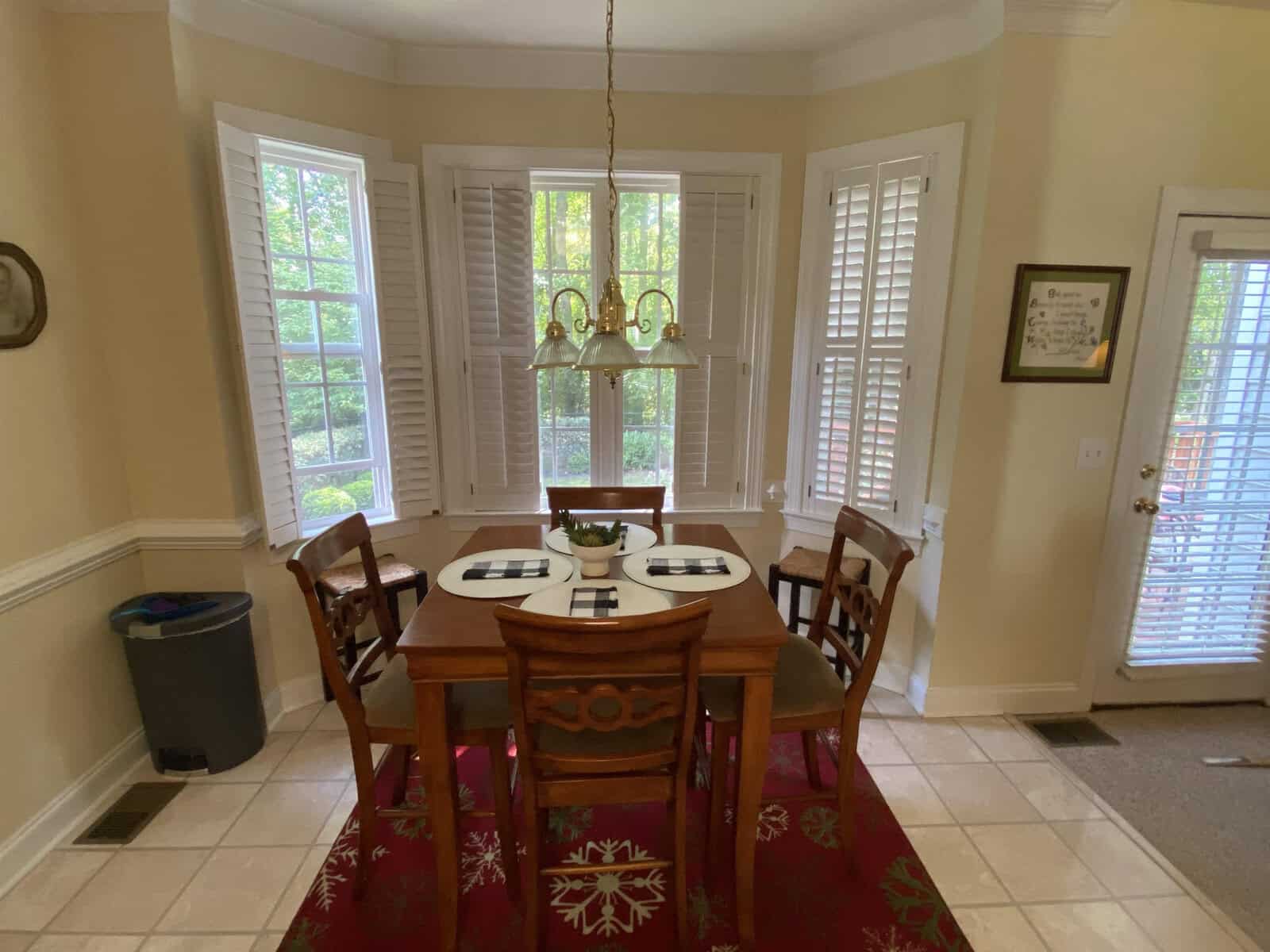 A well-lit dining area with a wooden table and chairs, chandelier overhead, bay windows with white shutters, and a red patterned area rug on tiled flooring.