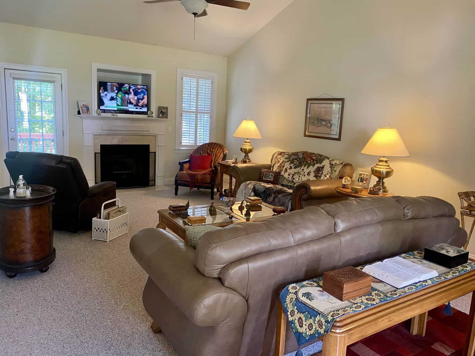 A cozy living room featuring a brown leather sofa, armchairs, a fireplace, and the television turned on, with warm lighting and visible windows.