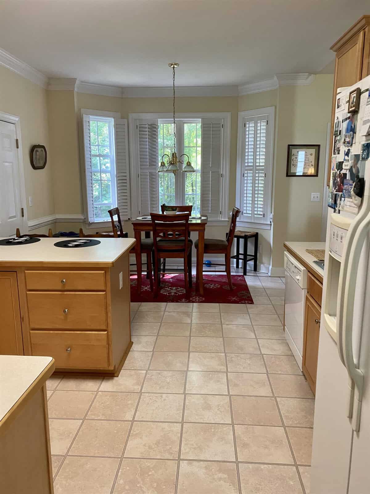A well-lit kitchen with tile flooring featuring a wooden dining table set on a red area rug, surrounded by bay windows with shutters, adjacent to a countertop with a kitchen sink and a refrigerator to the right. a wall with family photographs is visible on the right.