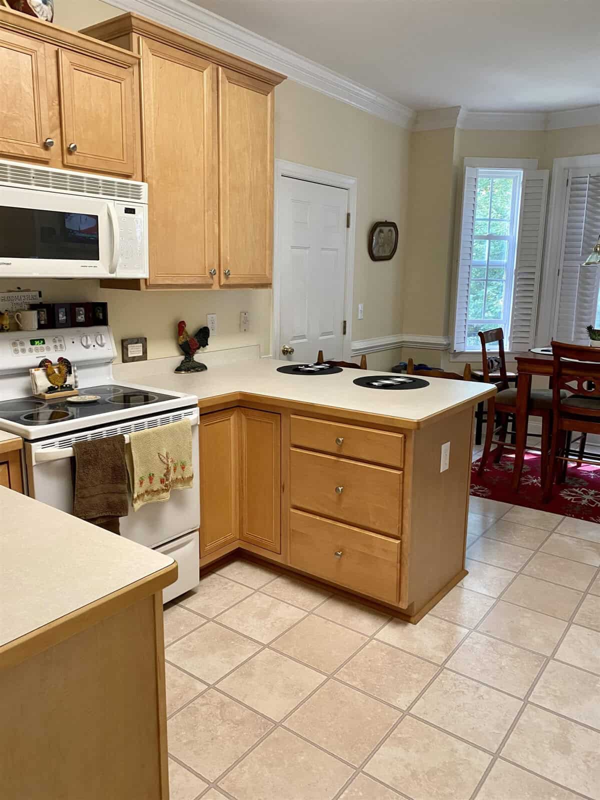 A clean, well-lit kitchen with wooden cabinetry, white countertops, modern appliances, and a dining area visible in the background.