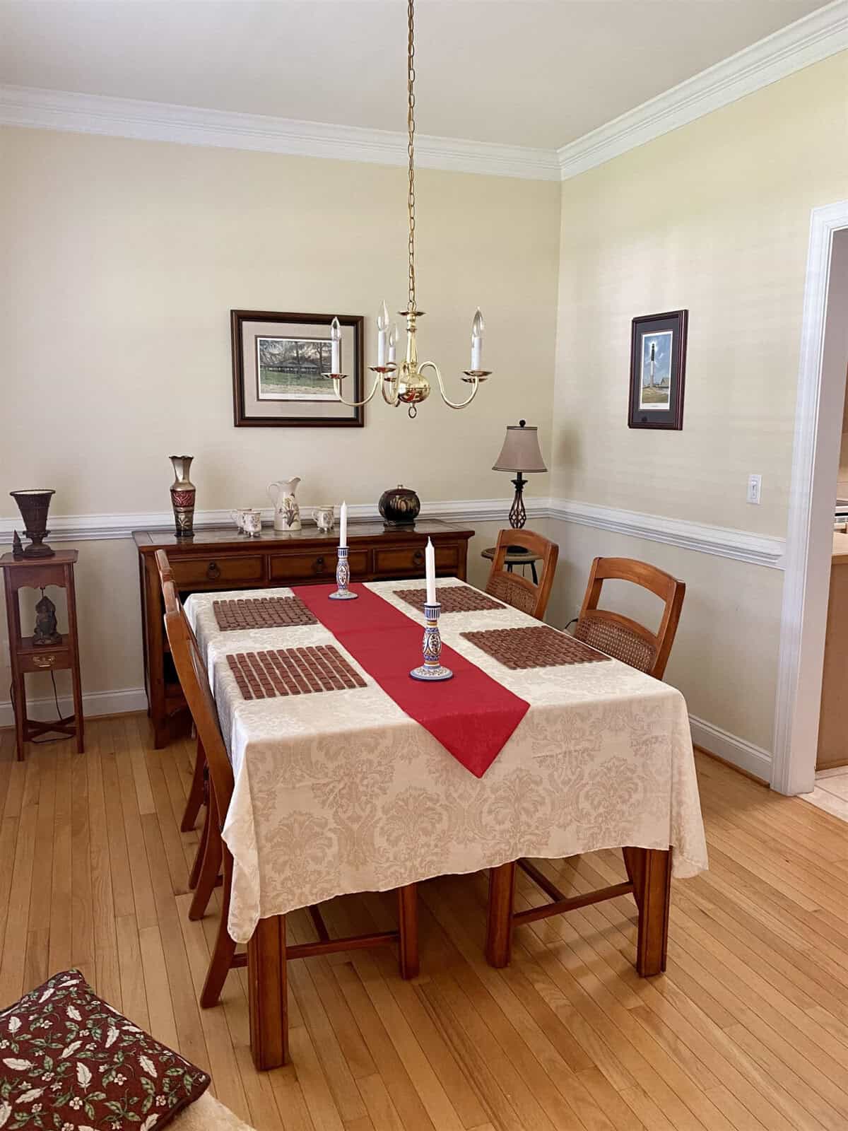 A well-appointed dining room featuring a wooden table set with placemats and a red runner, complemented by a matching chandelier above and decorative items on surrounding furniture.