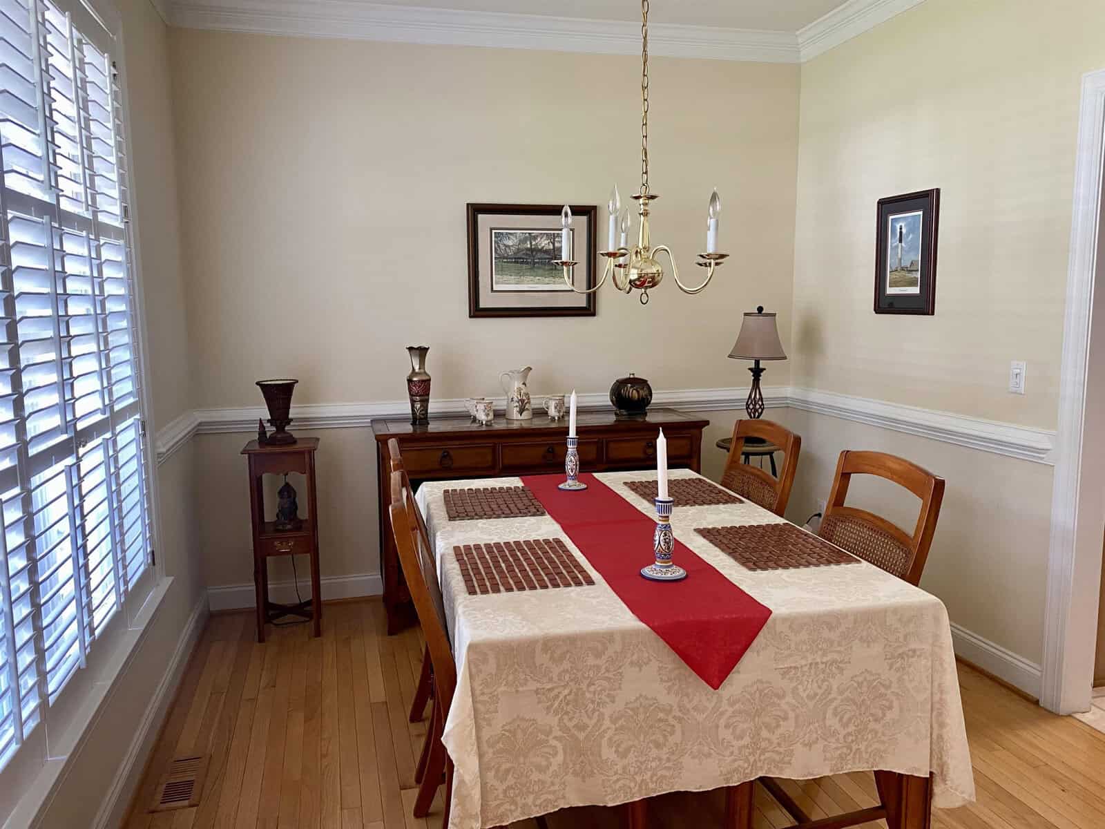 A neatly arranged dining room featuring a wooden table with a cream tablecloth and a red runner, surrounded by matching chairs, a chandelier above, and various decorative items including a lamp, vases, and framed artwork on the walls.