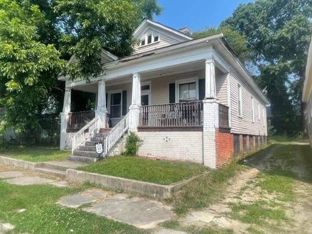 A weathered white house at 126 Zene Street, Henderson NC, with a front porch, featuring a set of stairs and a metal railing, situated on a grassy lot with some foliage in