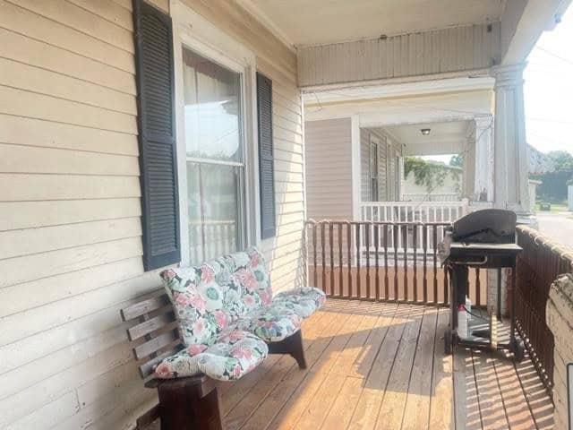 A wooden porch featuring a bench with floral cushions, black shutters on windows, and a barbecue grill to the right.