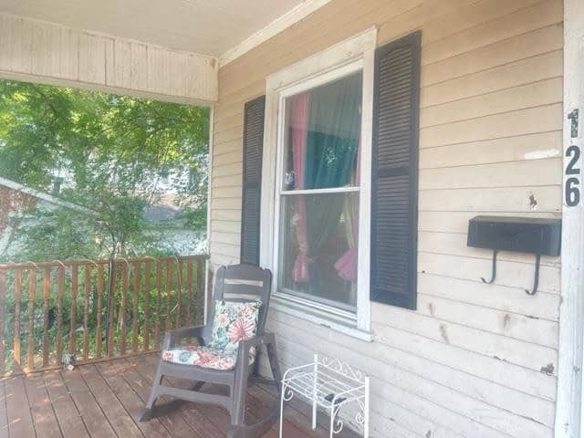 A wooden chair with a floral cushion and a small white table sit on a porch with beige siding, next to a large window with partially drawn curtains.