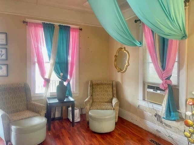 A vintage-styled room with hardwood floors, featuring two draped windows with multicolored curtains, a fireplace, and two upholstered armchairs with a small round table in between.
